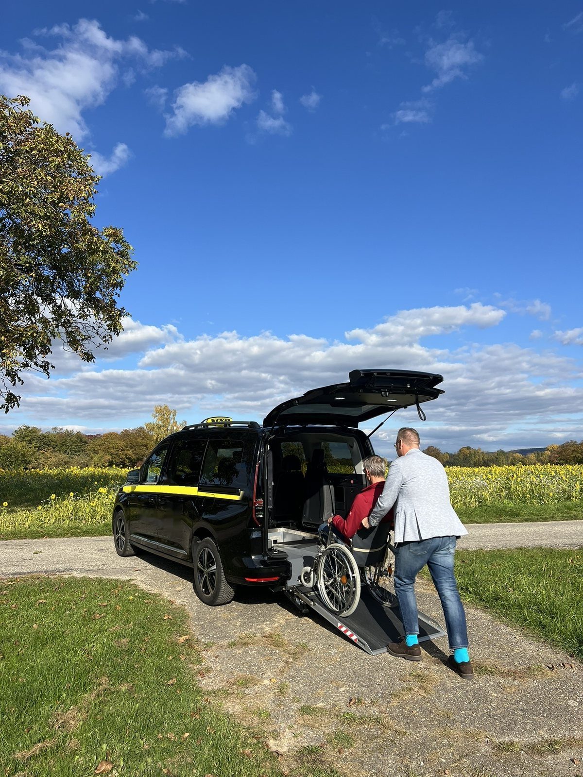 Ein Mann schiebt einen Fahrgast im Rollstuhl über eine Rampe in das Heck eines Taxis, im Hintergrund ein Sonnenblumenfeld