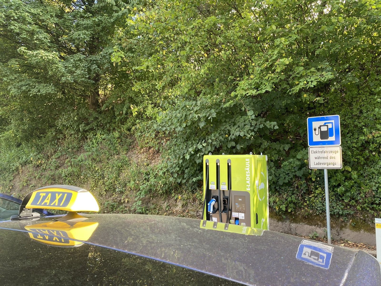 Taxi mit Dachschild steht an einer Schnellladesäule, daneben das Verkehrszeichen für Elektrofahrzeuge während des Ladevorgangs