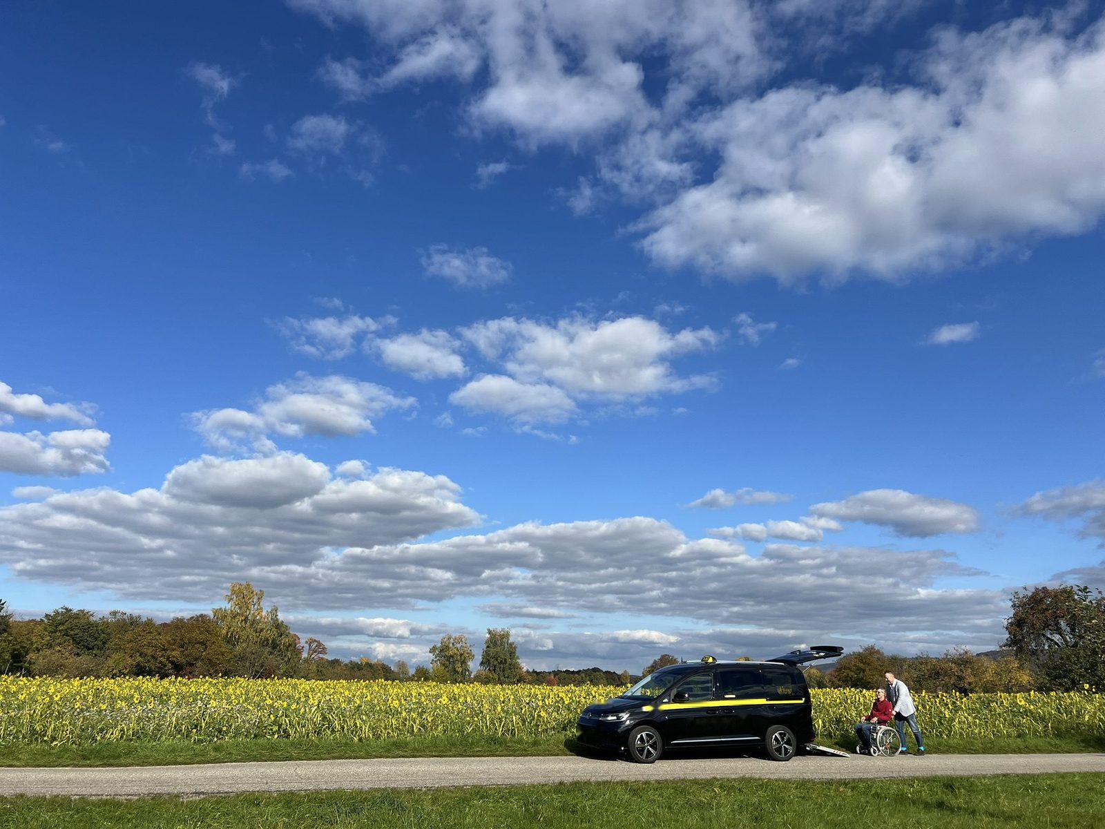 Taxi mit ausgeklappter Rollstuhlrampe vor einem Sonnenblumenfeld, daneben ein Fahrgast im Rollstuhl mit Begleitperson