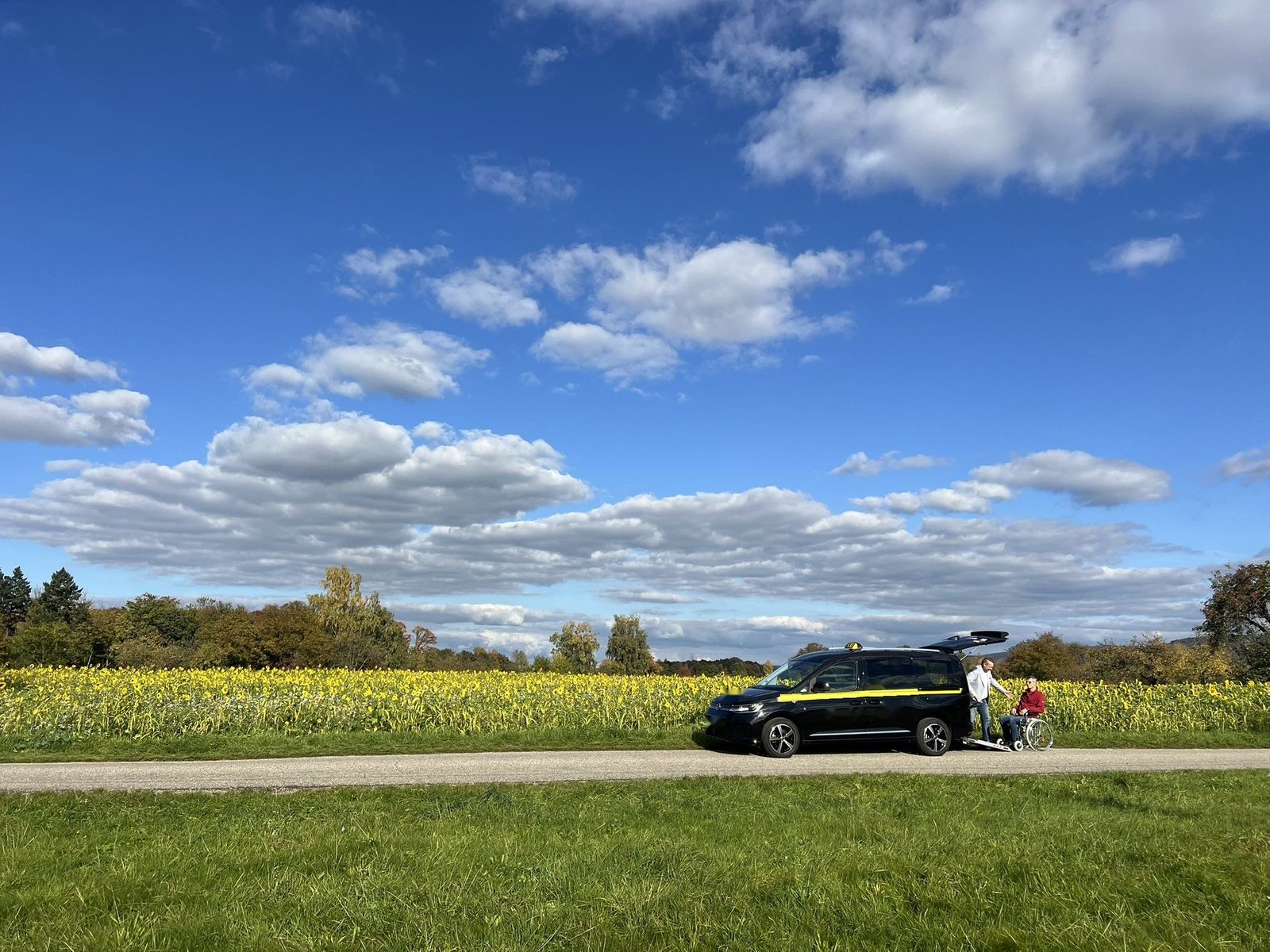 Taxi mit ausgeklappter Rampe auf einem Feldweg vor einem Sonnenblumenfeld, daneben ein Fahrgast im Rollstuhl und eine Begleitperson, darüber weiter blauer Himmel